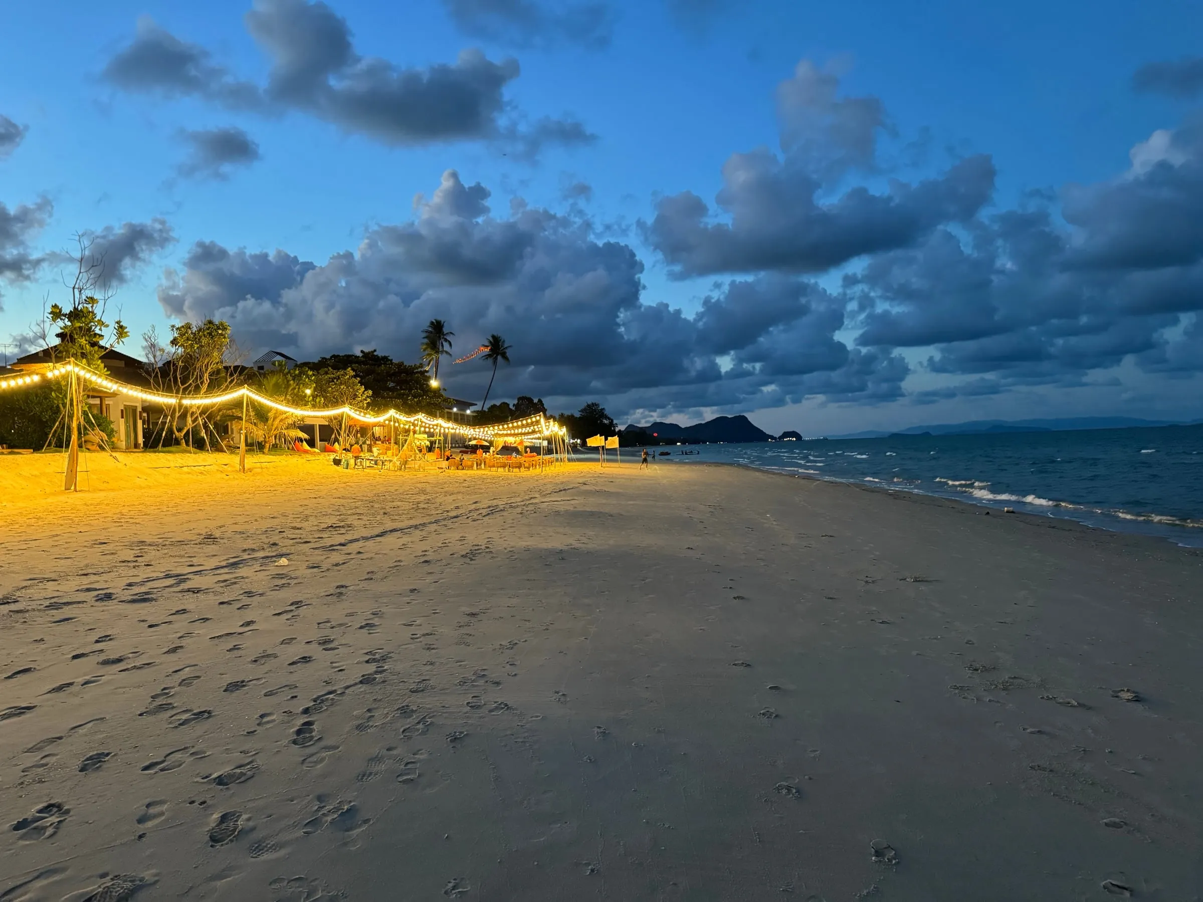 String lights over a beach bar at dusk, the sand wide and quiet toward the headland