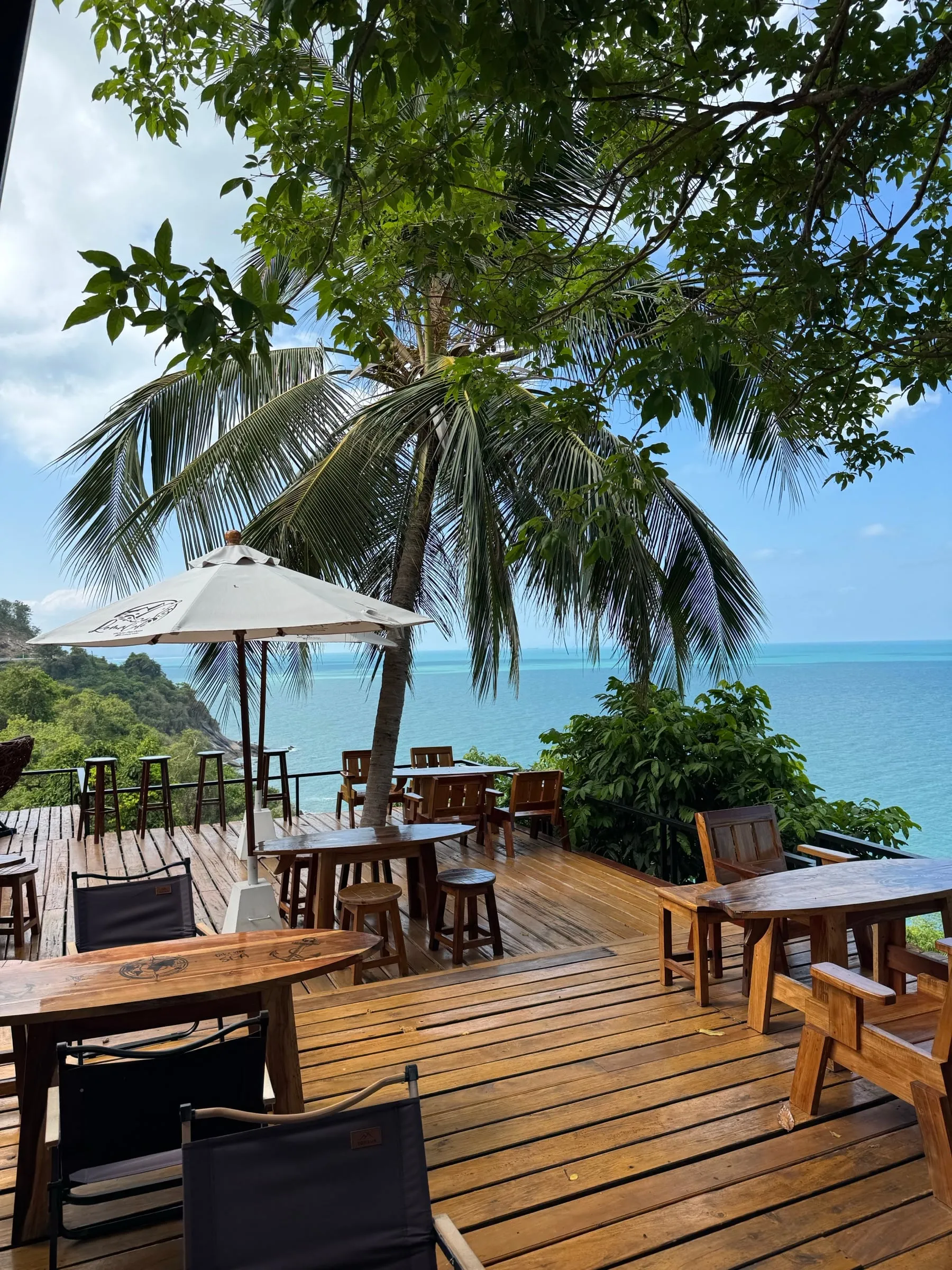 A wooden cafe deck on the hillside under a palm, looking out over the open sea